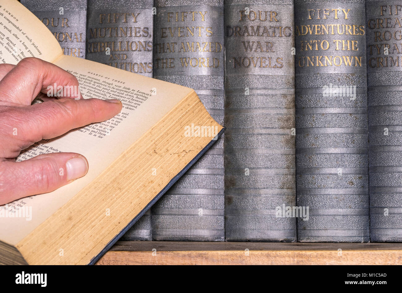 Man's hand on an open book, published in English, with a shelf of vintage hard back books in the background. England, UK. Stock Photo