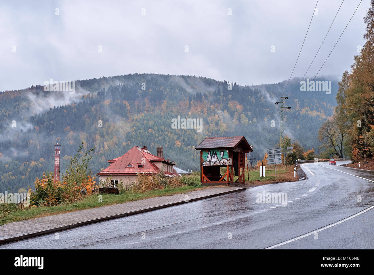 Fog and mist over the pine mountain forest with traditional old village houses, a red car on the road and a fancy bus stop Stock Photo