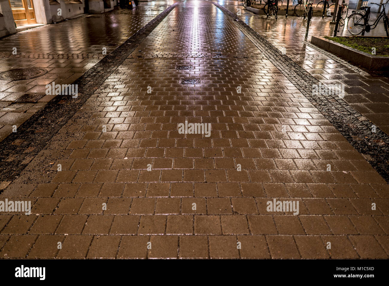 Rainy night over a pedestrian street with cobblestone paving and lights reflection from the nearby buildings and street illumination. Bicycles perked. Stock Photo