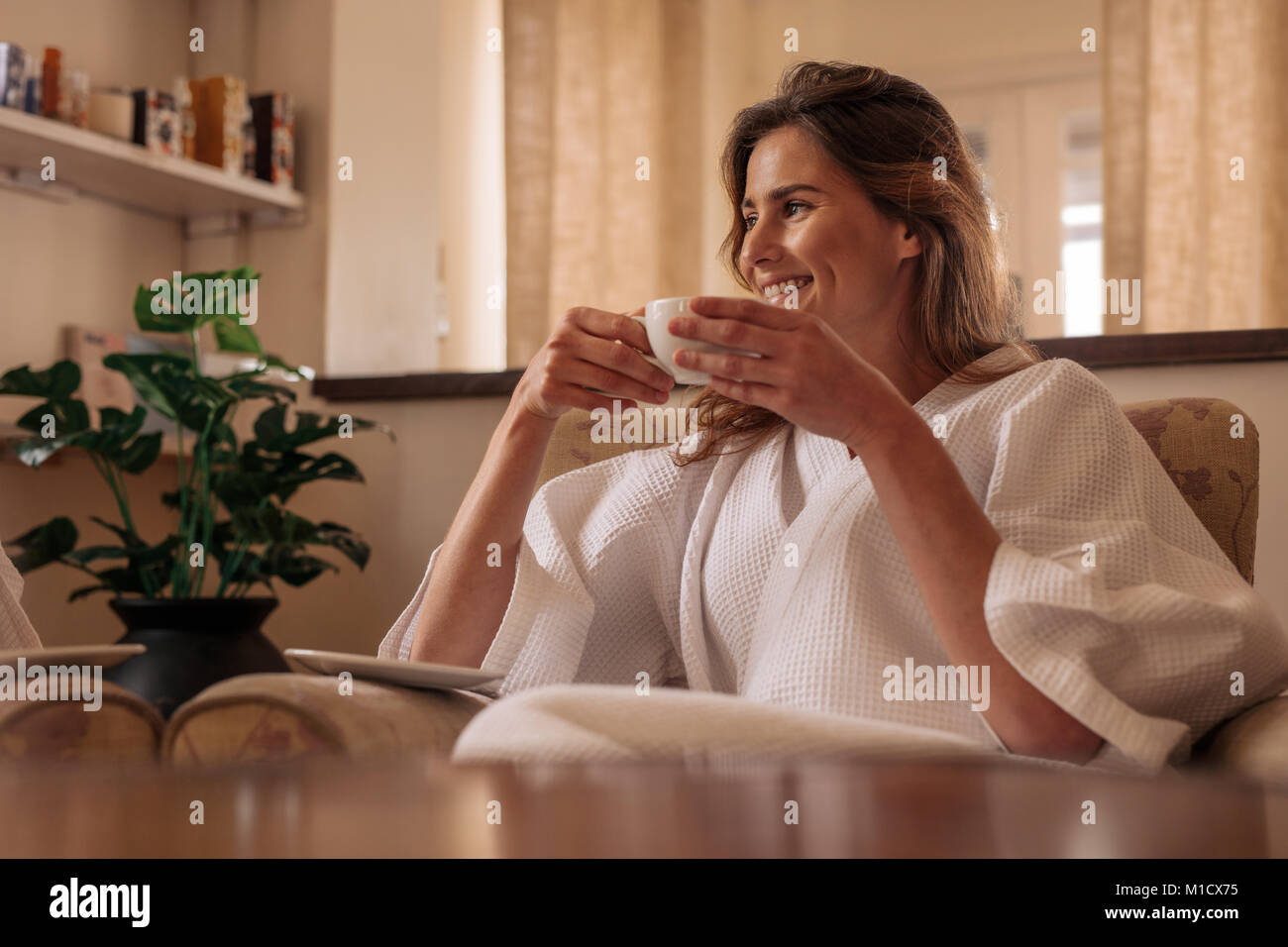 Smiling woman in bathrobe sitting on chair and drinking coffee at health spa. Female having coffee and relaxing at spa salon. Stock Photo