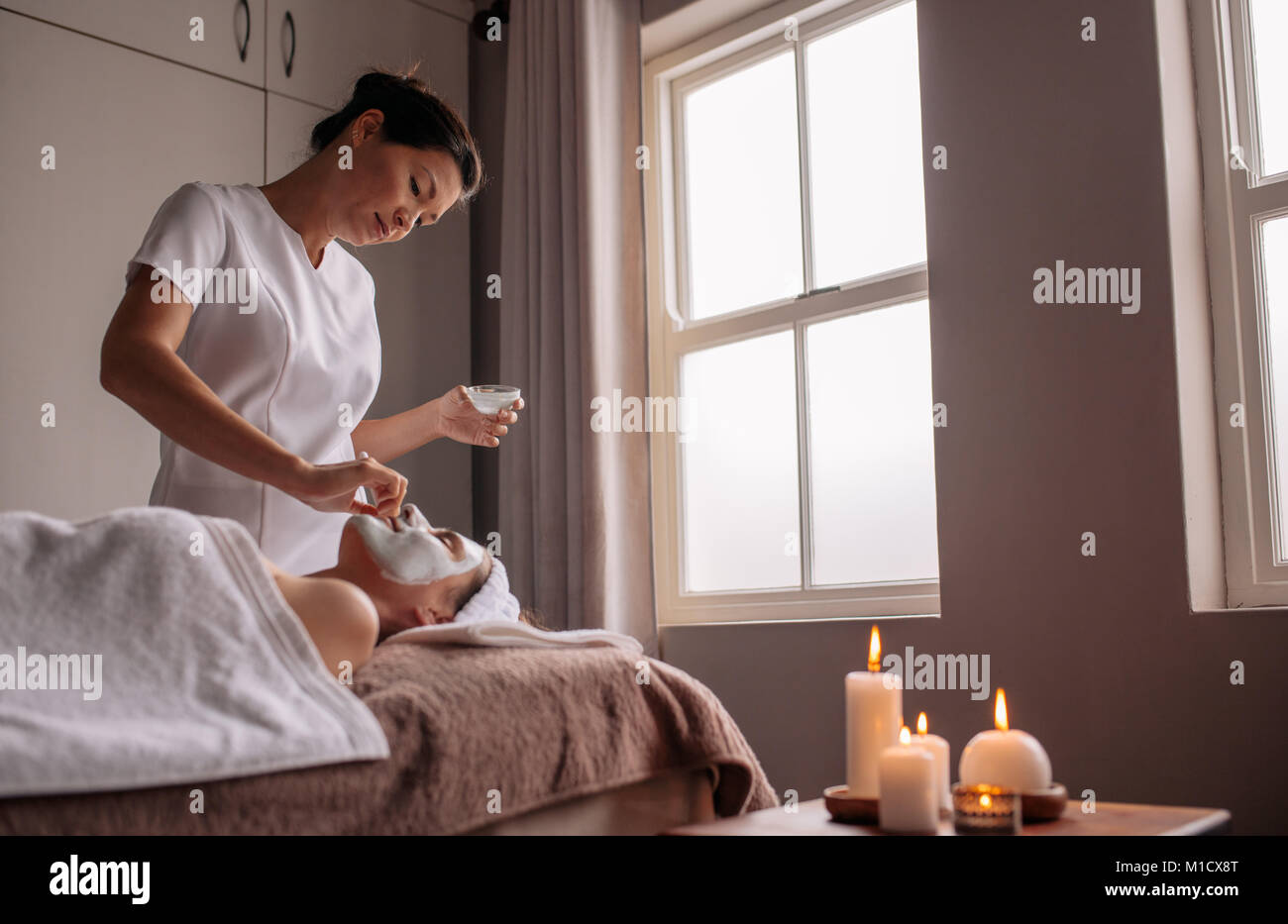 Woman receiving a face mask treatment in a beauty spa by therapist. Experienced cosmetician applying facial mask on female face. Stock Photo