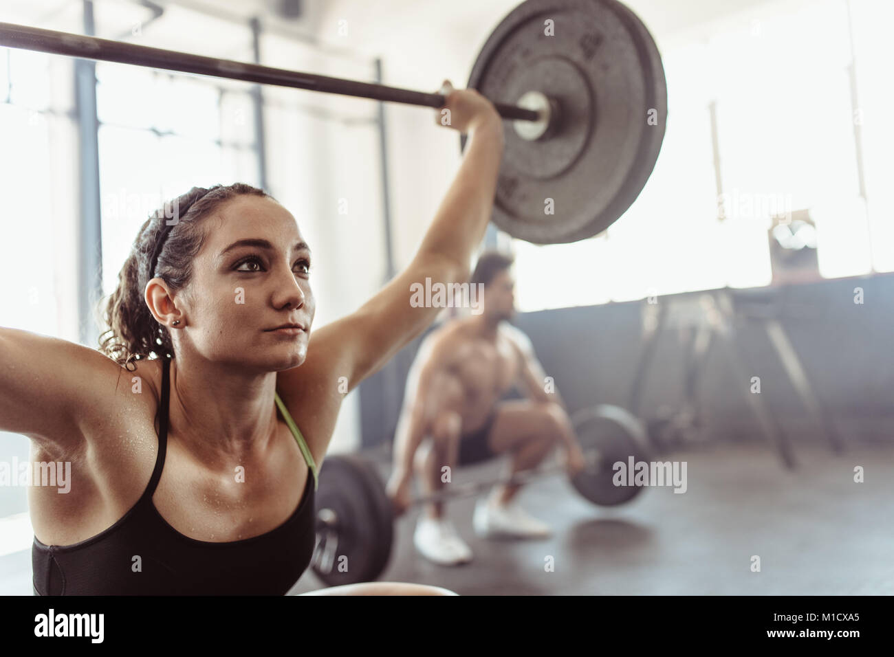 Tough young woman exercising with barbell. Determined female athlete lifting heavy weights at cross training gym. Stock Photo