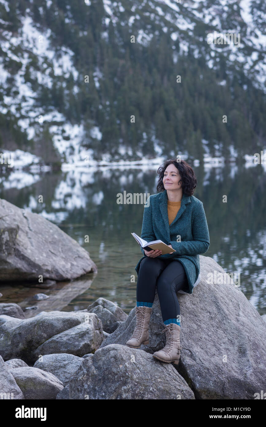 Thoughtful woman sitting with book at lakeside Stock Photo