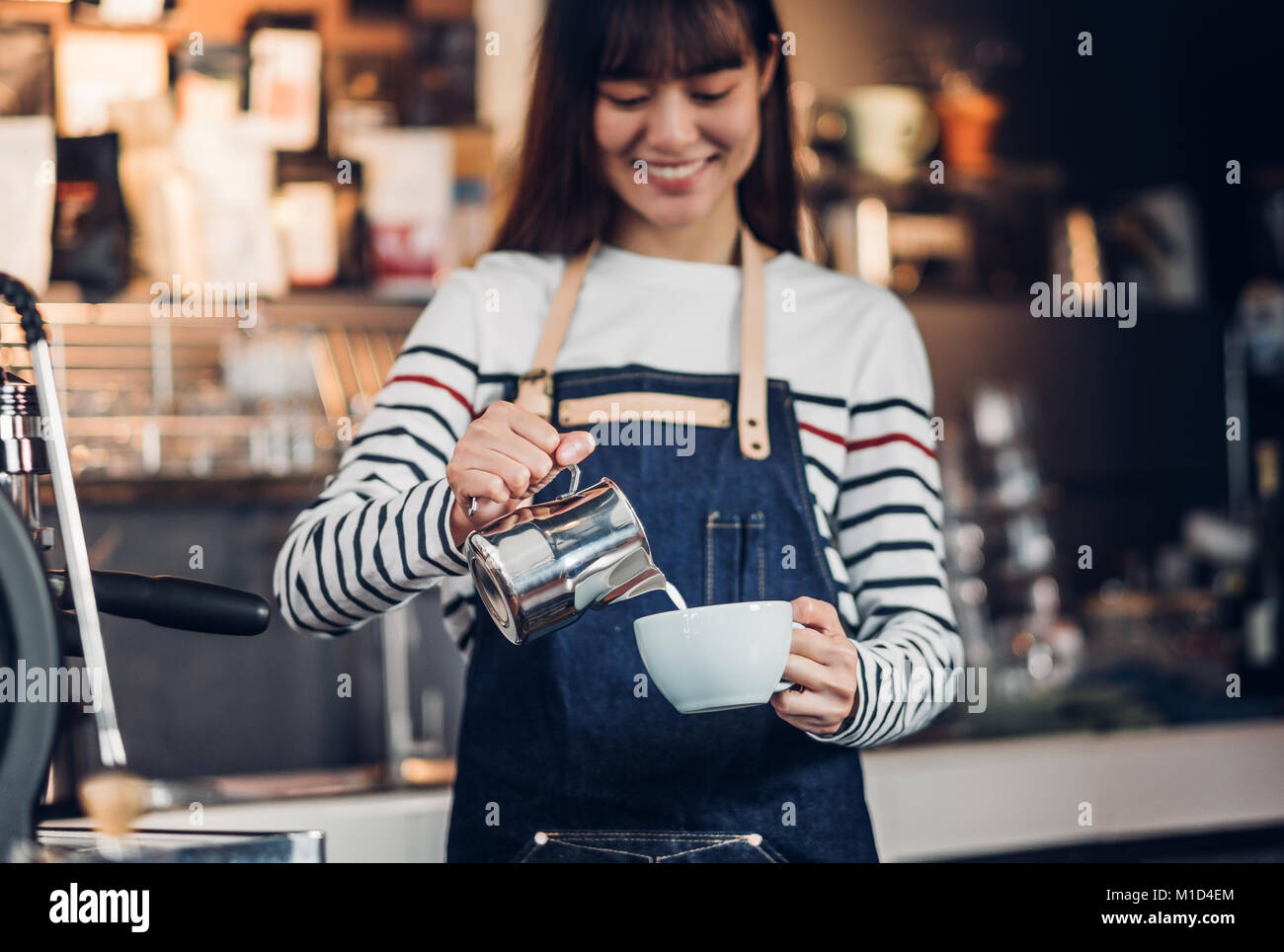 Asia woman barista pour milk into hot coffee cup at counter bar in front of machine in cafe restaurant,Food business owner concept Stock Photo