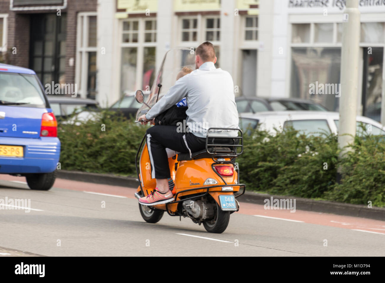 Under Dutch law it is not a requirement for anyone owing a motorbike or scooter under 30cc to wear a crash helmet.   A father riding his scooter with  Stock Photo