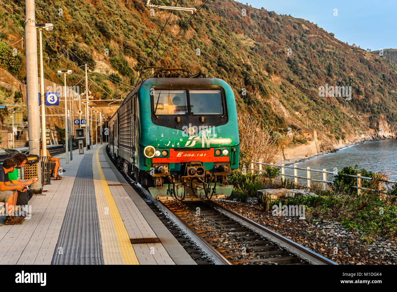 A train travels into the Monterosso al Mare train station on the Cinque Terre coast of Italy as travelers sit and enjoy a warm day in early autumn Stock Photo