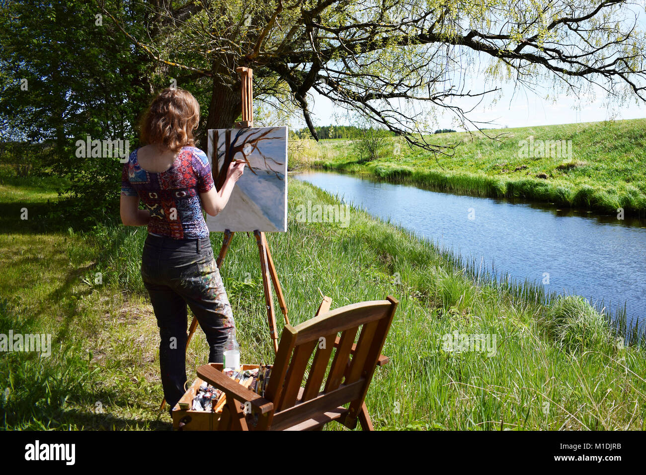 Woman artist paints landscape painting of small river. Painter rear view. Stock Photo