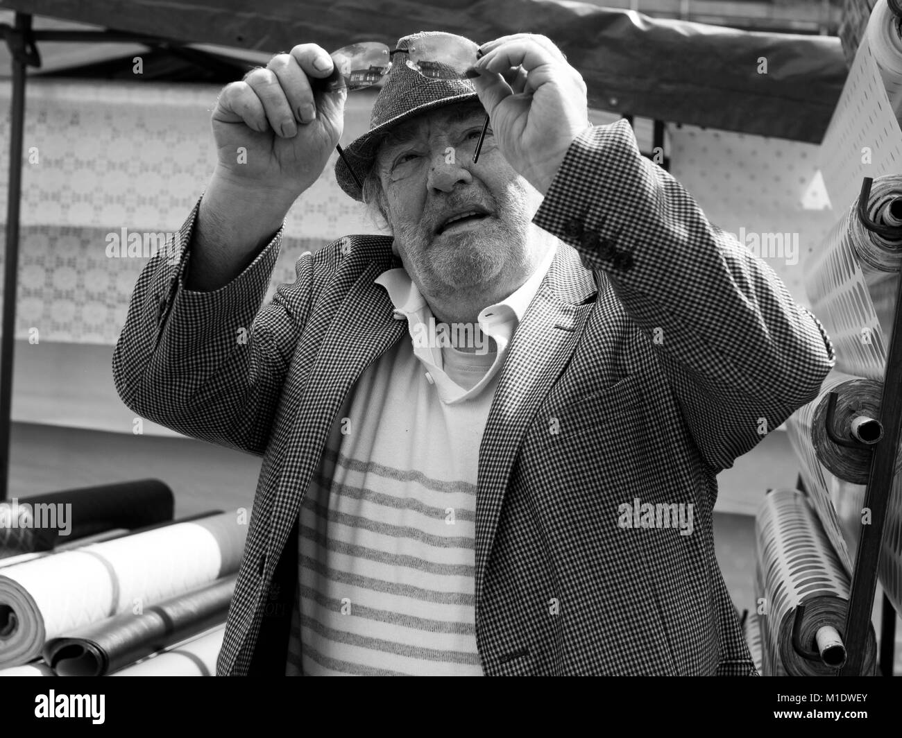 Market trader wearing tweed jacket and hat in front of rolls of fabric looks up at his glasses - Monochrome image Stock Photo