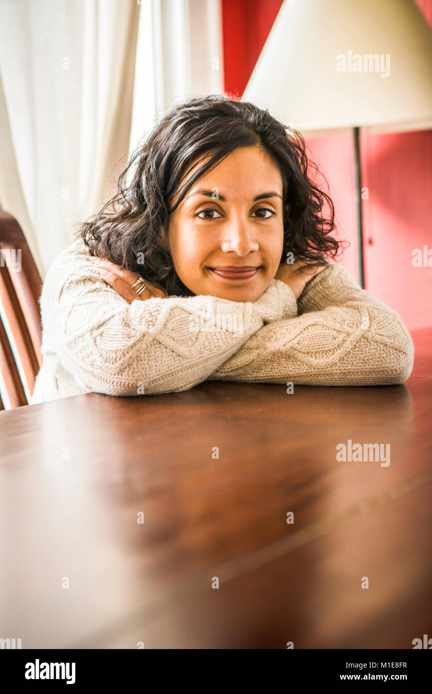 A portrait of a smiling early 30's Sri Lanken - American woman leaning on a table and resting her head on her arms. Stock Photo