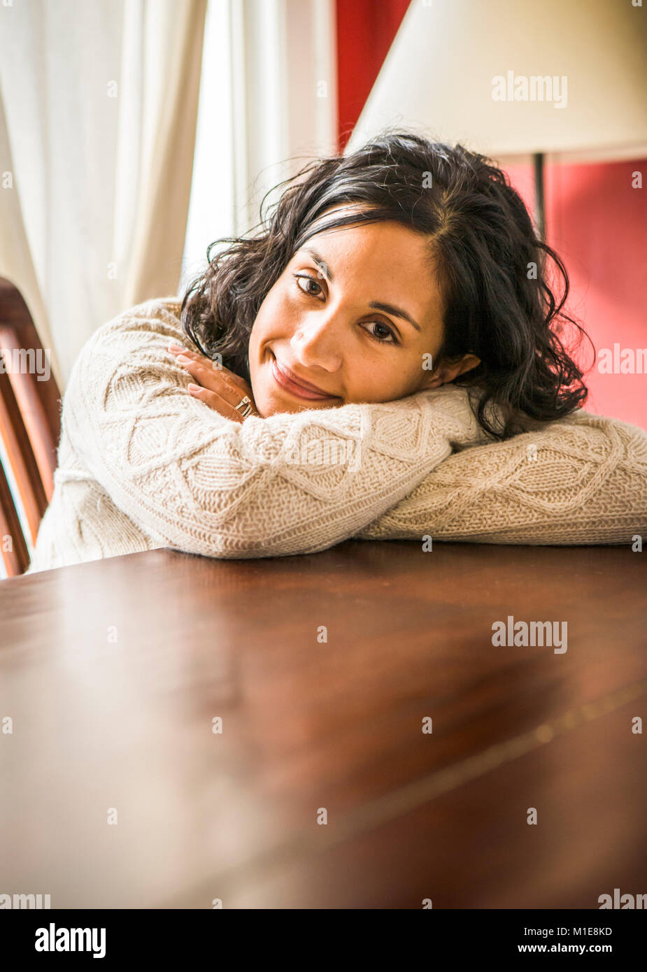 A portrait of a smiling early 30's Sri Lanken - American woman leaning on a table and resting her head on her arms. Stock Photo