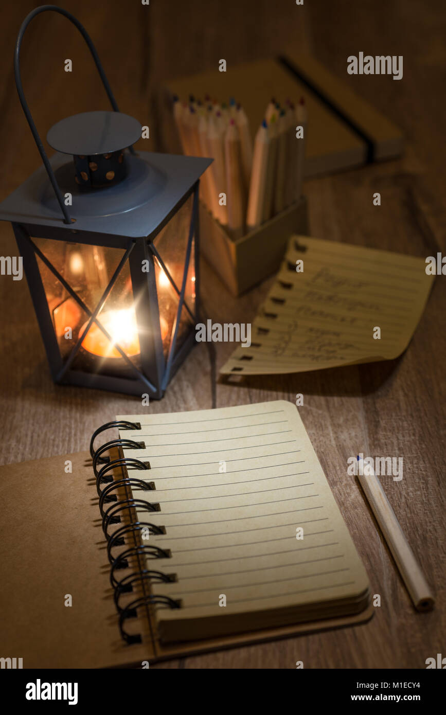 Open notebook with blank front page, candle in metal vintage lantern, torn-out page and a pencil on wooden table. Space for your notes, recipe or a po Stock Photo
