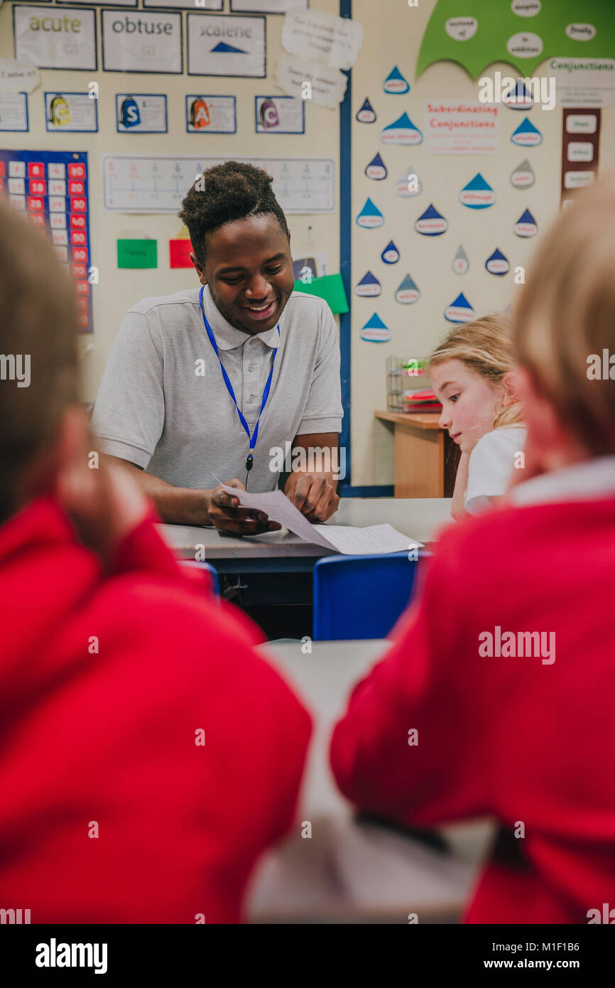 Young teacher is working with his primary school students in the classroom. He is holding a sheet of paper and is discussing it happily with the stude Stock Photo