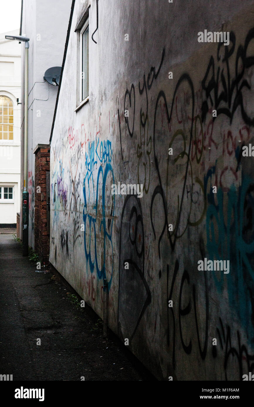 The side of a white painted house seen down the side of an old alley way covered in graffiti, words, pictures adn writing. Stock Photo