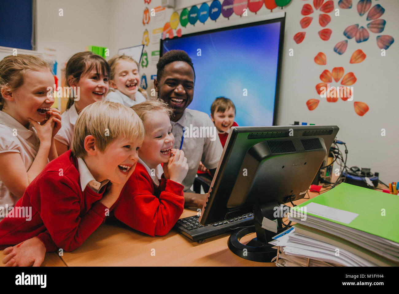 Group of primary school students are crowded round a computer with their teacher. They are all laughing and looking at the computer screen. Stock Photo