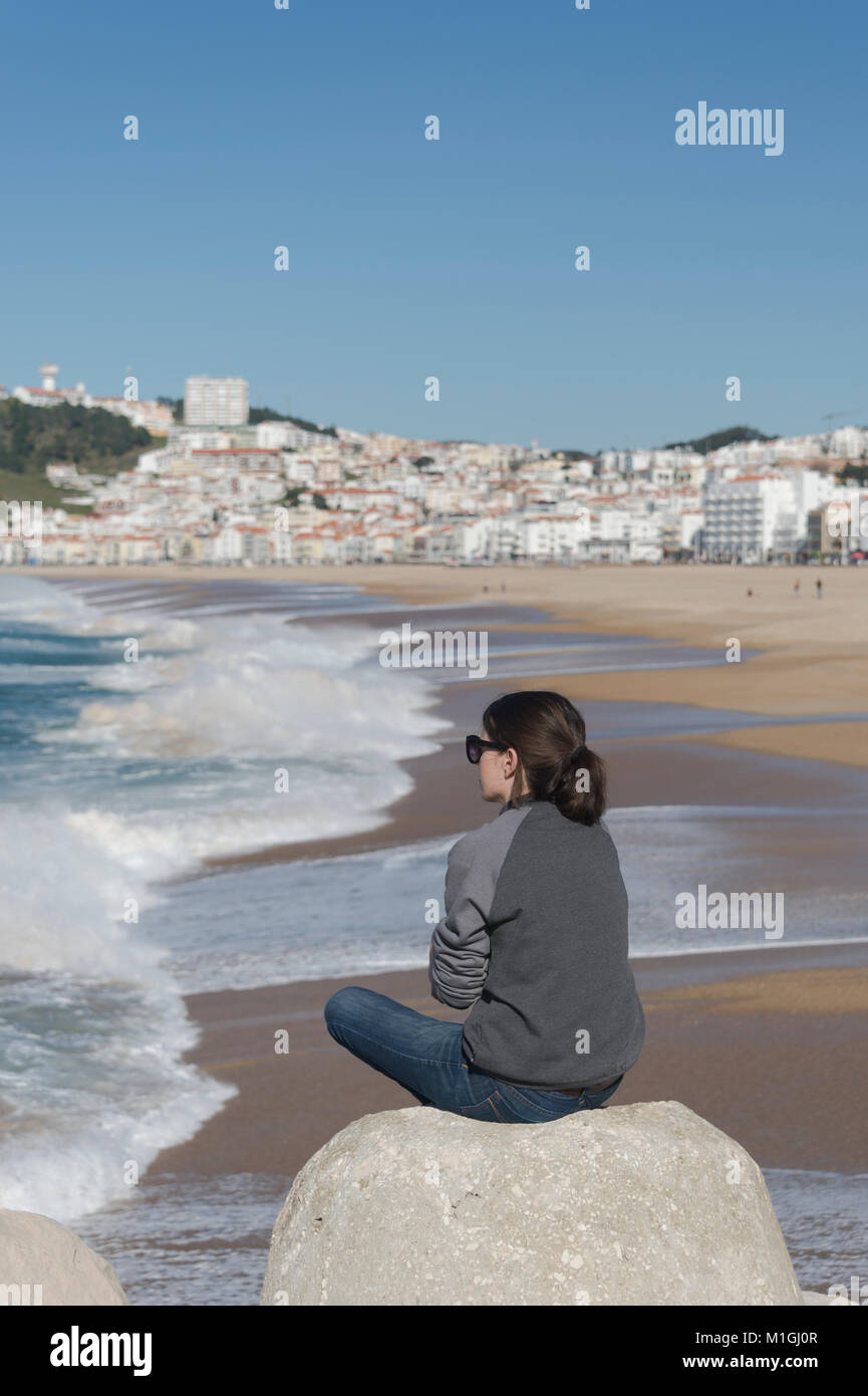 woman sitting on a rock watching the waves at Nazare, Portugal. Stock Photo