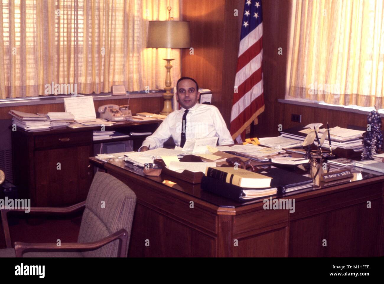 Photograph of Dr. David Sencer, CDC (Centers for Disease Control) director (1966-1977), sitting behind a desk covered with papers, an American flag visible behind him, 1970. Image courtesy CDC. () Stock Photo