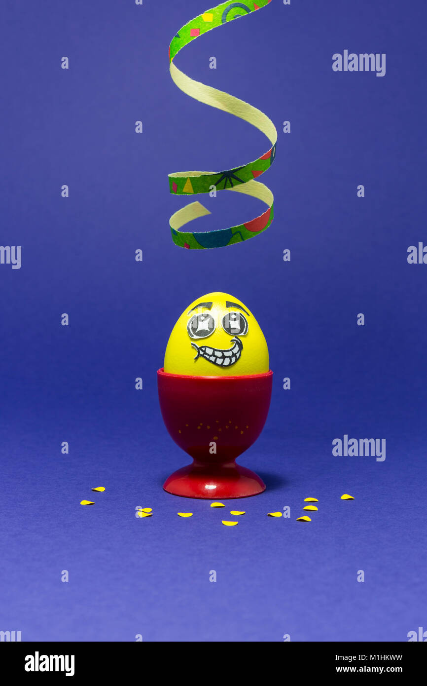 Yellow painted Easter egg with funny cartoon style face in a red plastic egg cup, colorful paper streamer and confetti on purple background Stock Photo