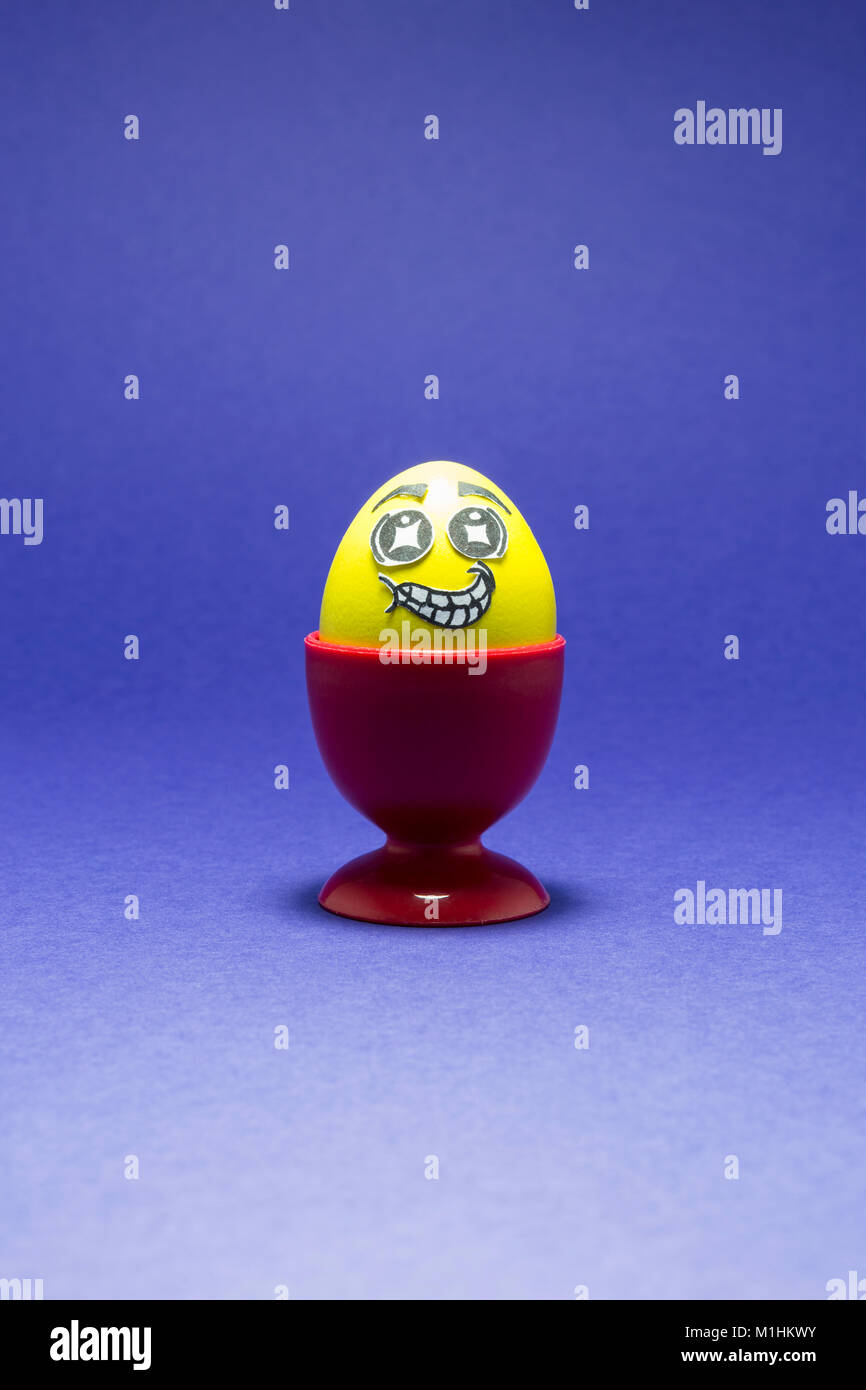 Yellow painted Easter egg with funny cartoon style face in a red plastic egg cup and purple background Stock Photo