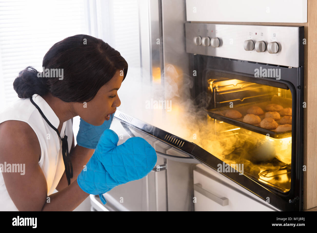 Shocked Woman Looking At Burnt Cookies With Smoke Coming From Oven