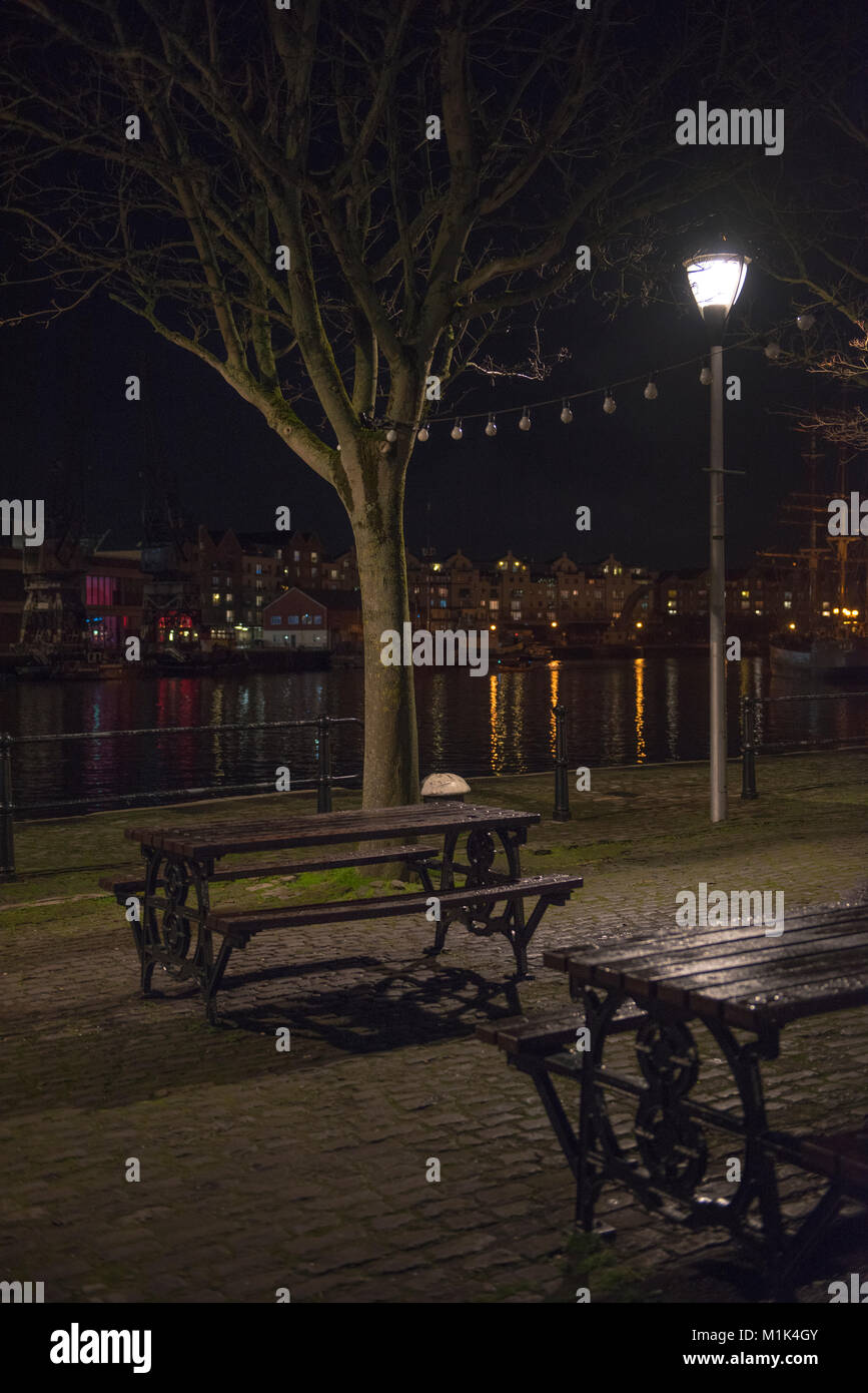 A night view of the Bristol harbour from the quay near Arnolfini, includes two picnic tables, a tree and a lamp. Stock Photo