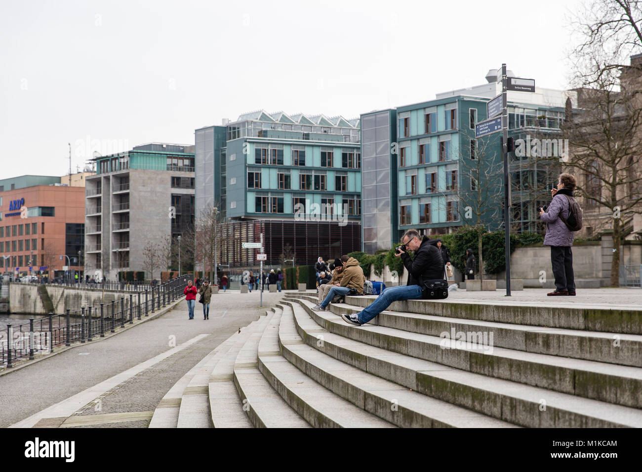 Man sitting on the steps in front of the river Spree in Berlin taking photographs of the modern architecture of the government district in Berlin Stock Photo