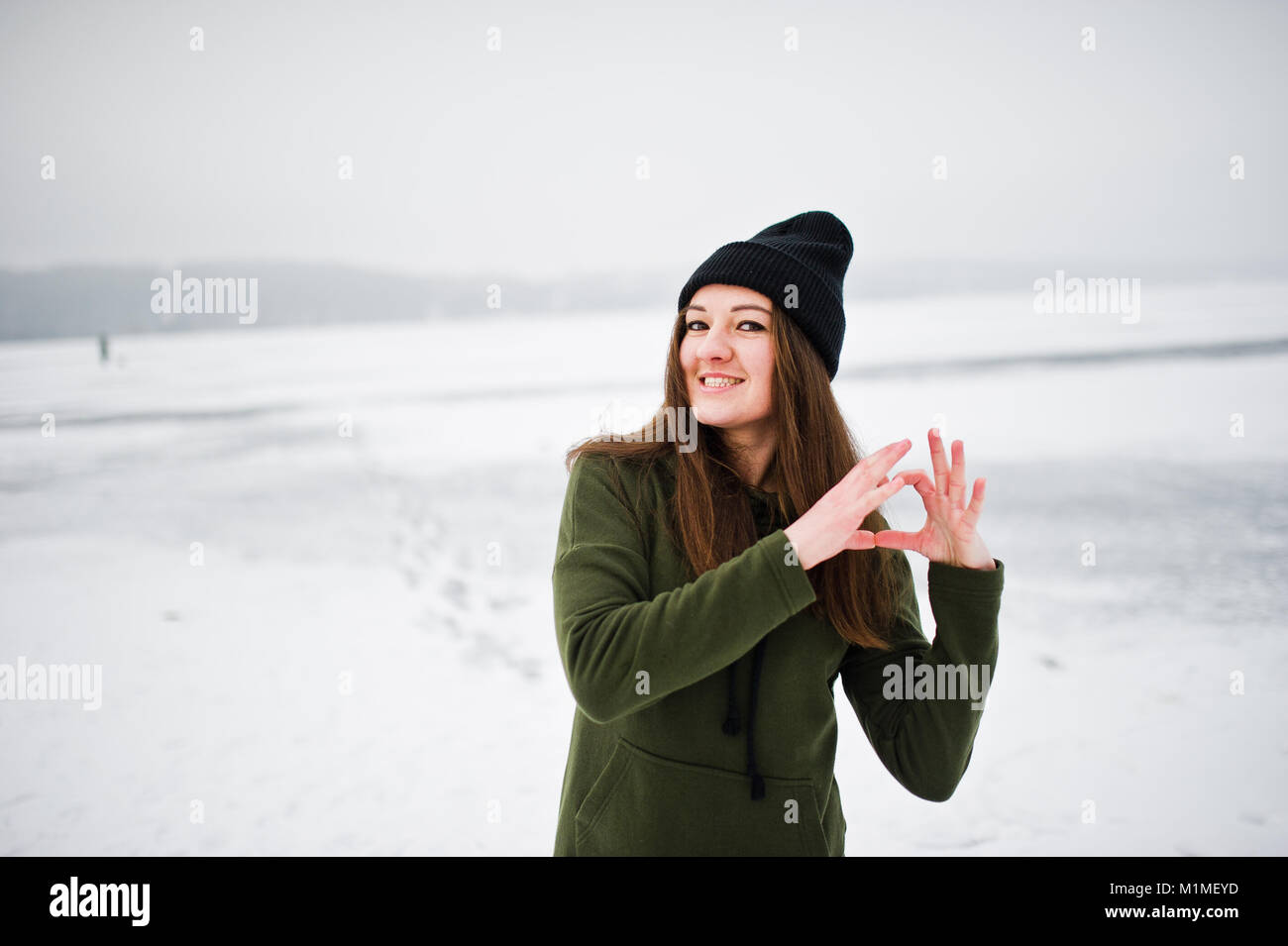 Funny girl wear on long green sweatshirt, jeans and black headwear, at frozen lake in winter day, showing heart on fingers. Stock Photo