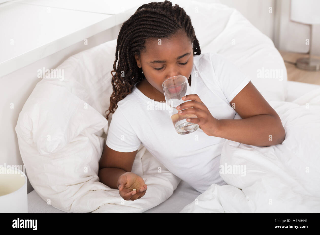 Sick African Girl In Bed Drinking Water While Taking Pill Stock Photo