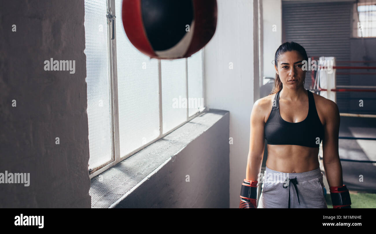 Female boxer standing inside a boxing studio. Woman boxer in her boxing gloves standing near a boxing speed bag. Stock Photo