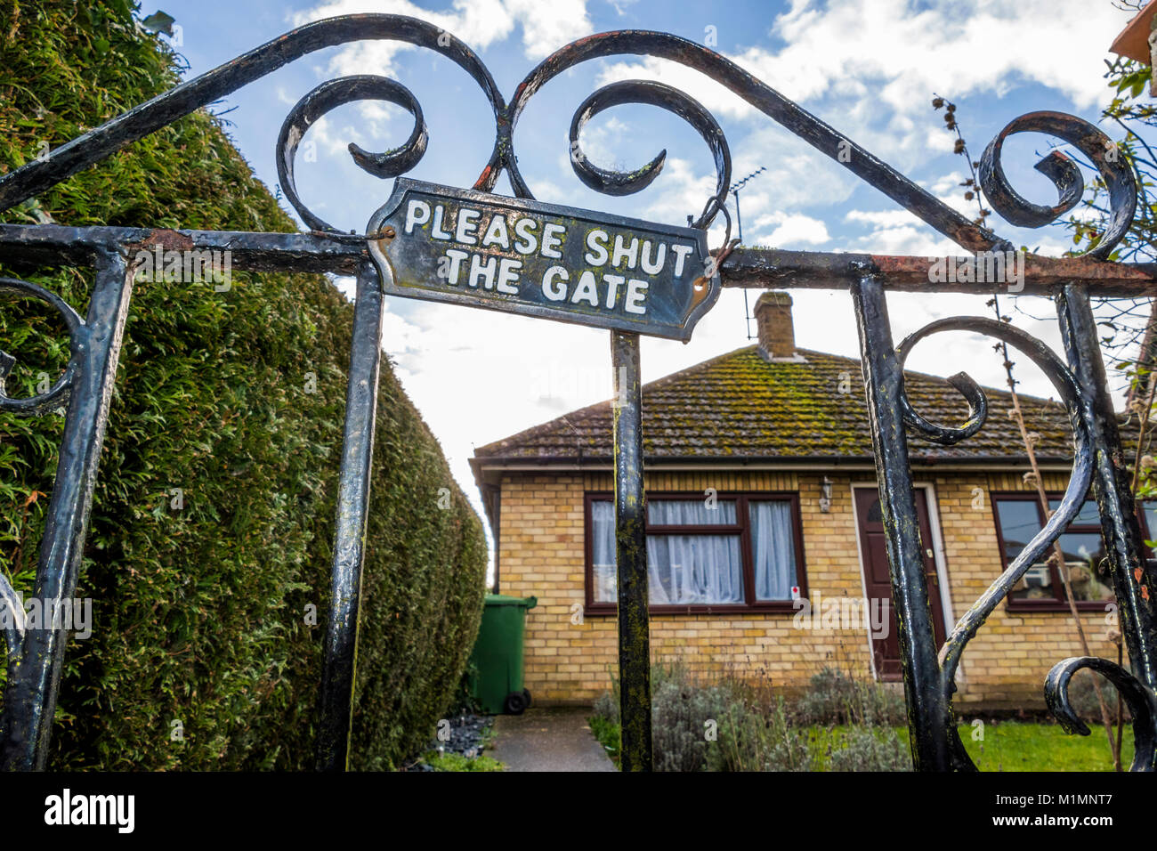 ‘Please shut the gate’ sign / notice attached to a wrought iron, rusty gate, next to the garden hedge of a 1960s single-storey bungalow. England, UK. Stock Photo