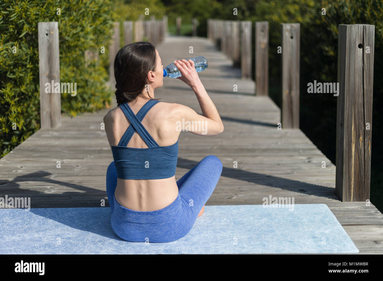 woman sitting on a yoga exercise mat drinking water from a bottle. Outdoors. Stock Photo