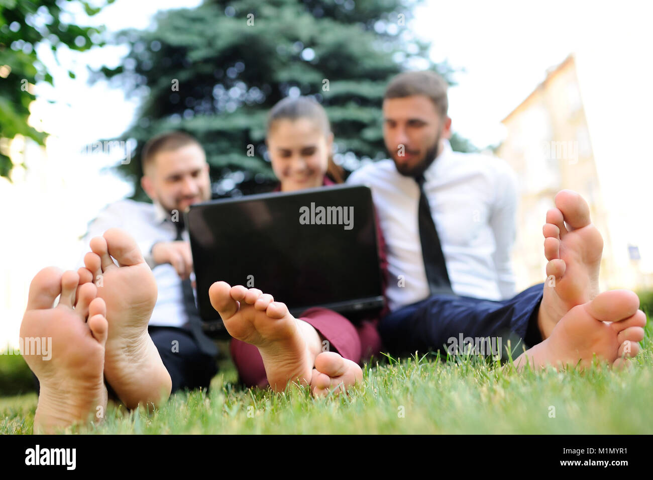 friends in business clothes and barefoot sitting on grass holding laptop in hands Stock Photo