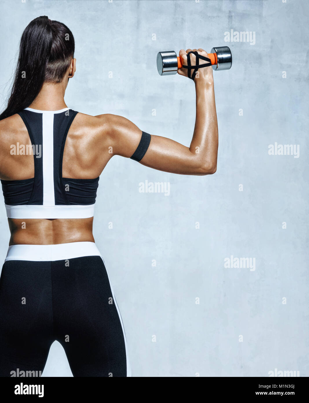 Strong woman working out with dumbbells, flexing her arm. Photo of sporty woman in sportswear on grey background. Rear view Stock Photo