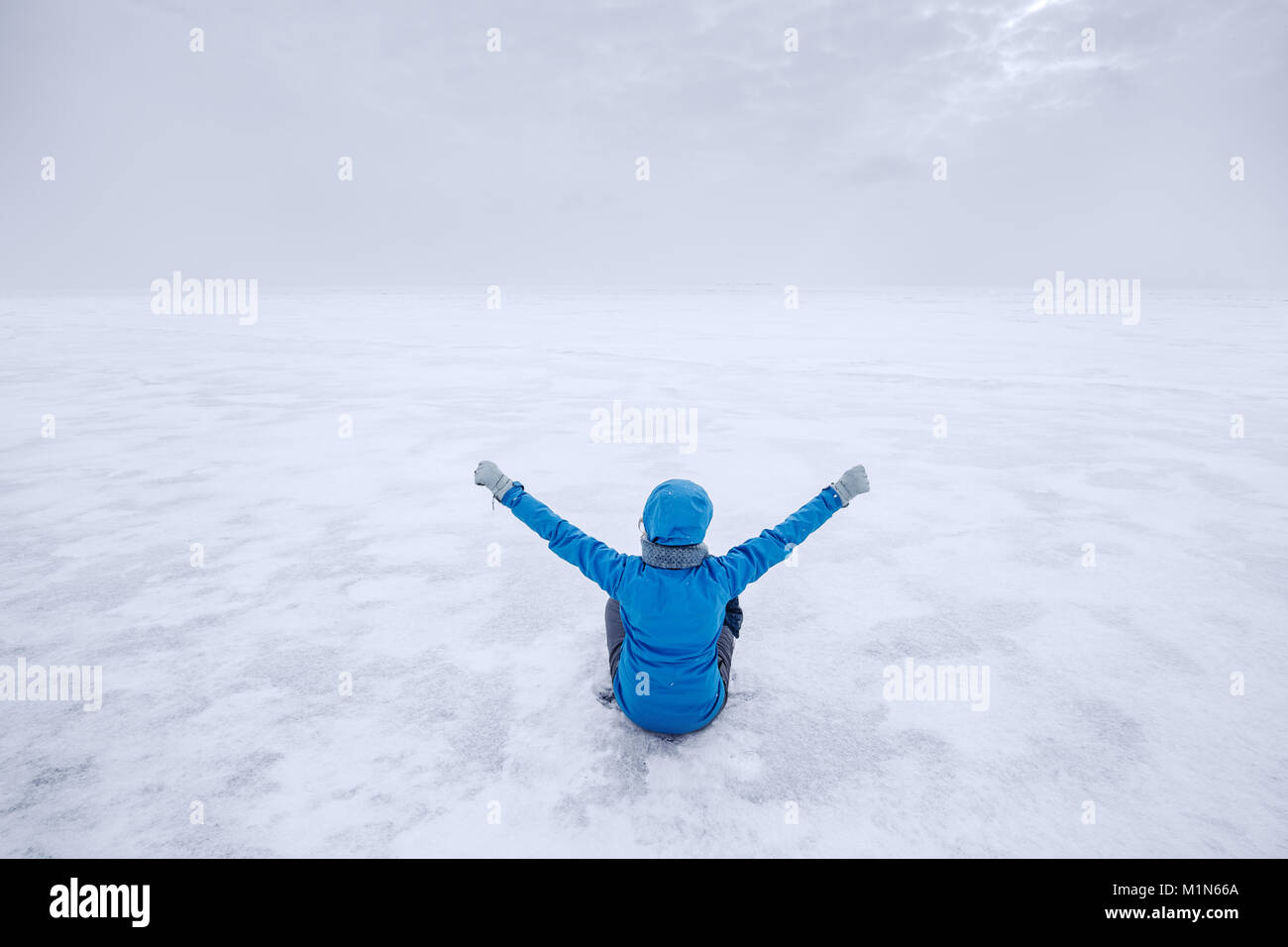 Woman in blue warm jacket sitting alone on the lake ice and staring forward. Cold cloudy winter day Stock Photo