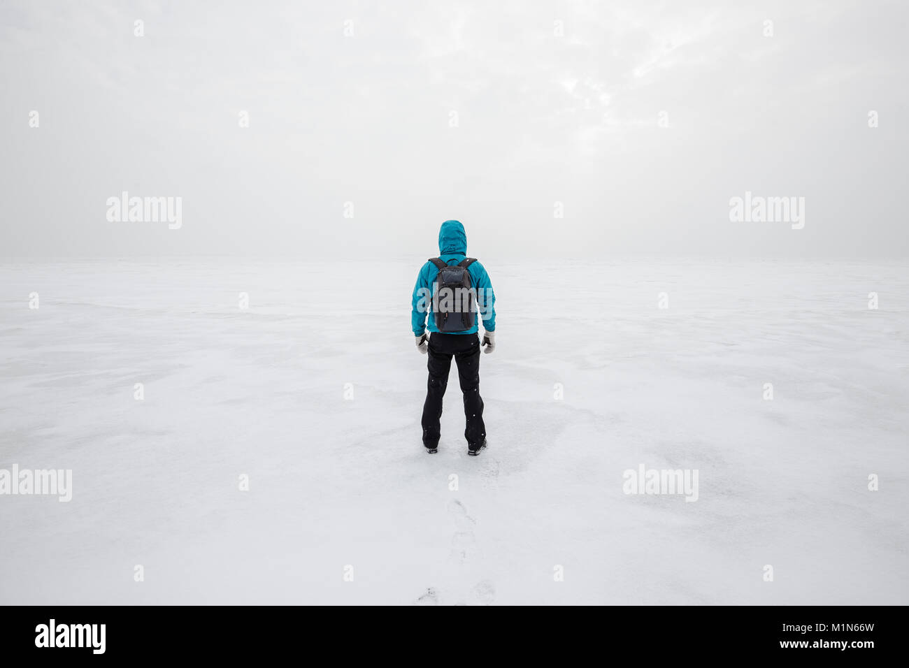 Man with backpack standing alone on the lake ice and staring forward. Cold cloudy winter day Stock Photo