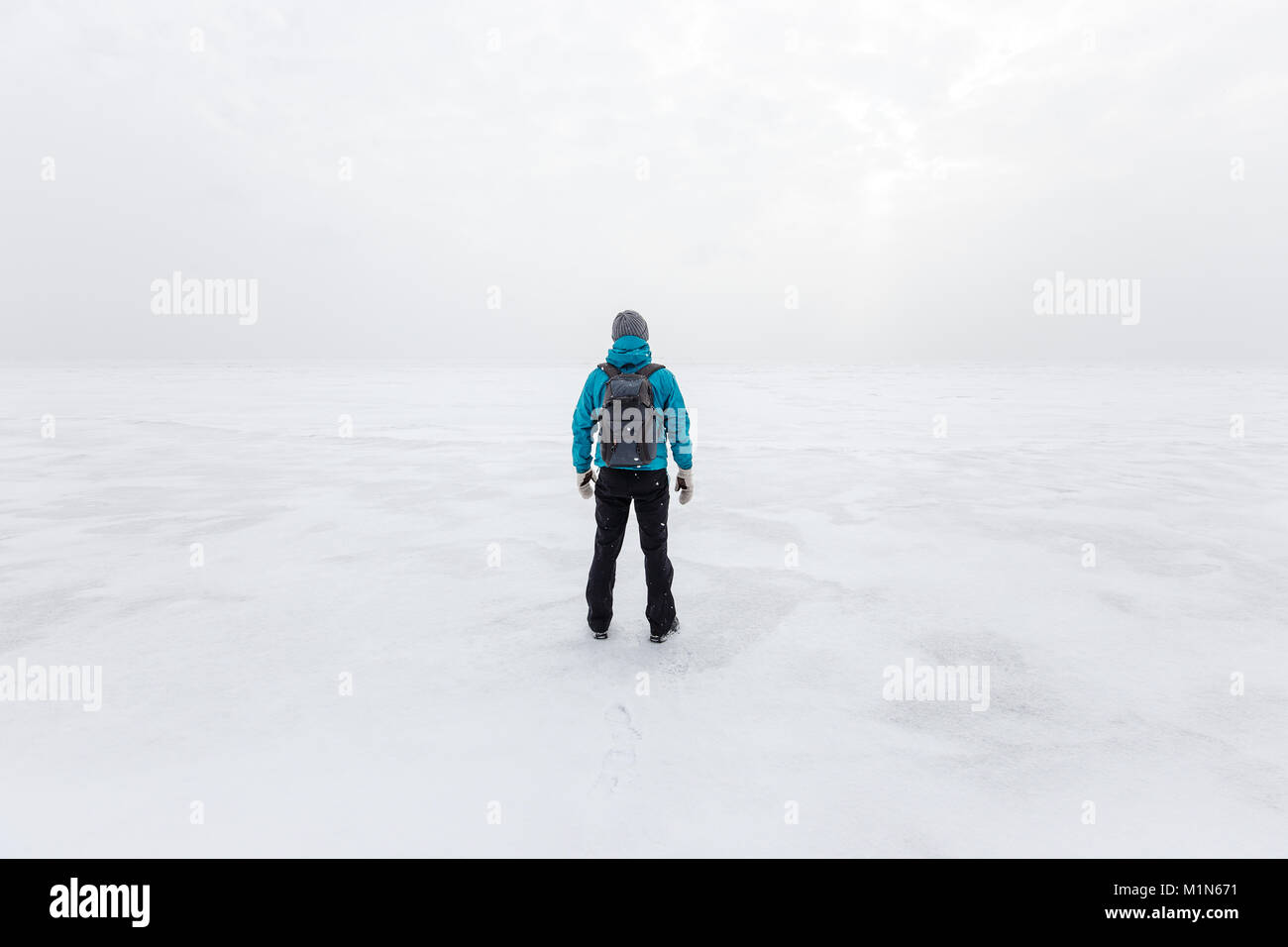 Man with backpack standing alone on the lake ice and staring forward. Cold cloudy winter day Stock Photo
