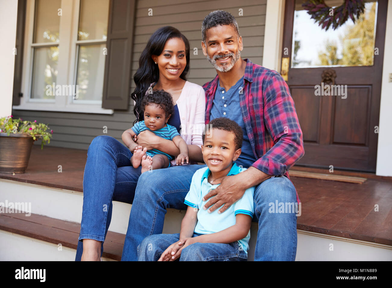 Family With Baby Son Sit On Steps Leading Up To Porch Of Home Stock Photo