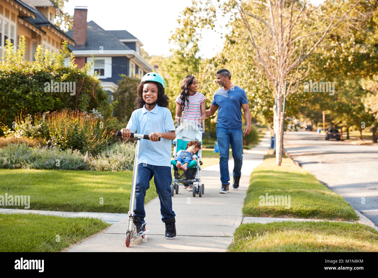 Couple Push Daughter In Stroller As Son Rides Scooter Stock Photo