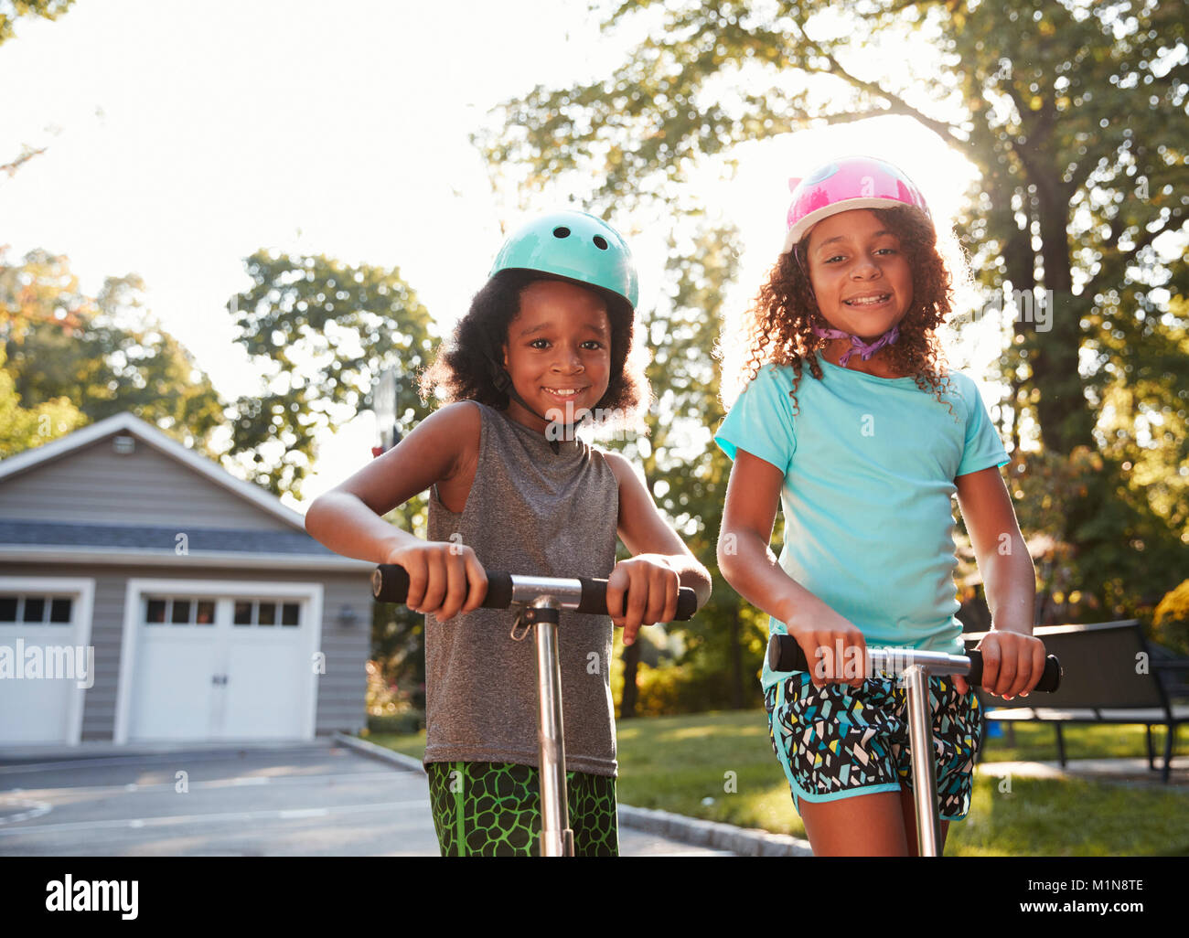 Brother And Sister Riding Scooters On Driveway At Home Stock Photo