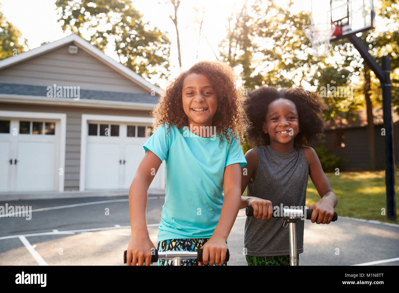 Brother And Sister Riding Scooters On Driveway At Home Stock Photo