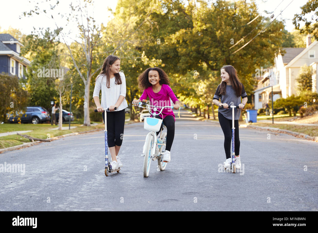 Three pre-teen girls riding in street on scooters and a bike Stock Photo