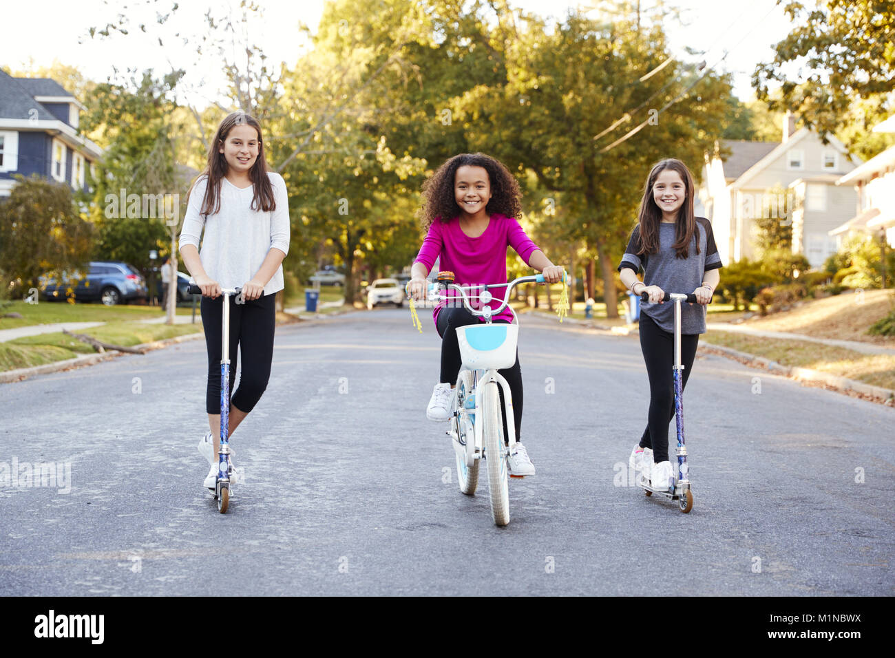 Three pre-teen girls on scooters and bike looking to camera Stock Photo