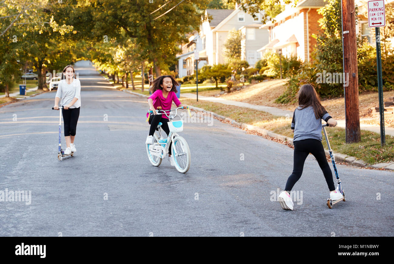 Three girls riding on scooters and a bike in the street Stock Photo