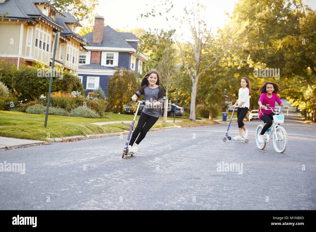 Three pre-teen girls playing in street on scooters and bike Stock Photo