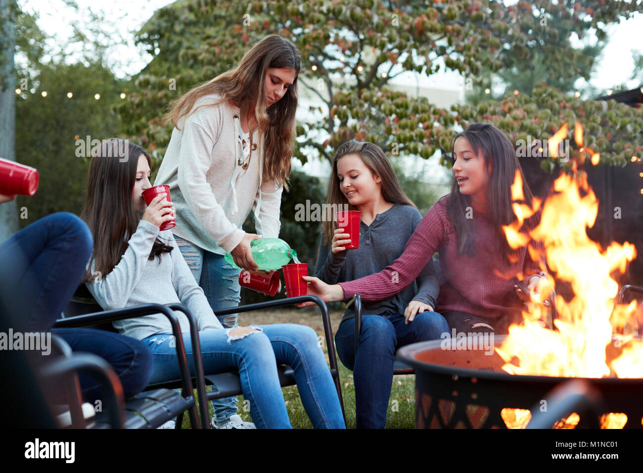 Teenager pours drinks for girlfriends sitting round firepit Stock Photo