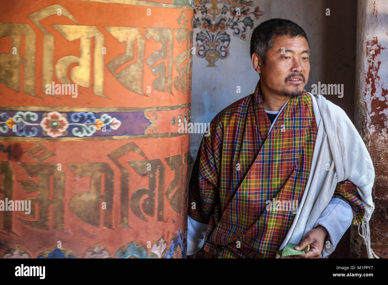 Bumthang, Bhutan.  Bhutanese Man Wearing Traditional Gho Turning Large Prayer Wheel.  Jambay Lhakhang Monastery. Stock Photo