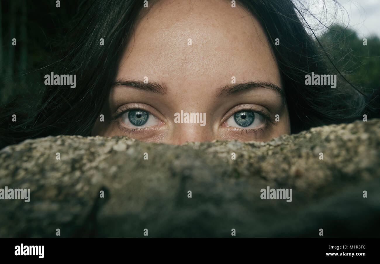 Chica de ojos azules y grandes mirando a camara por encima de una piedra tapandose la mitad de la cara Stock Photo