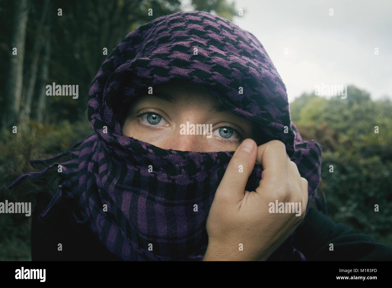 Chica de ojos azules mirando a camara tapandose la cabeza y parte de la cara con un pañuelo aguantado por su mano en la naturaleza. Stock Photo