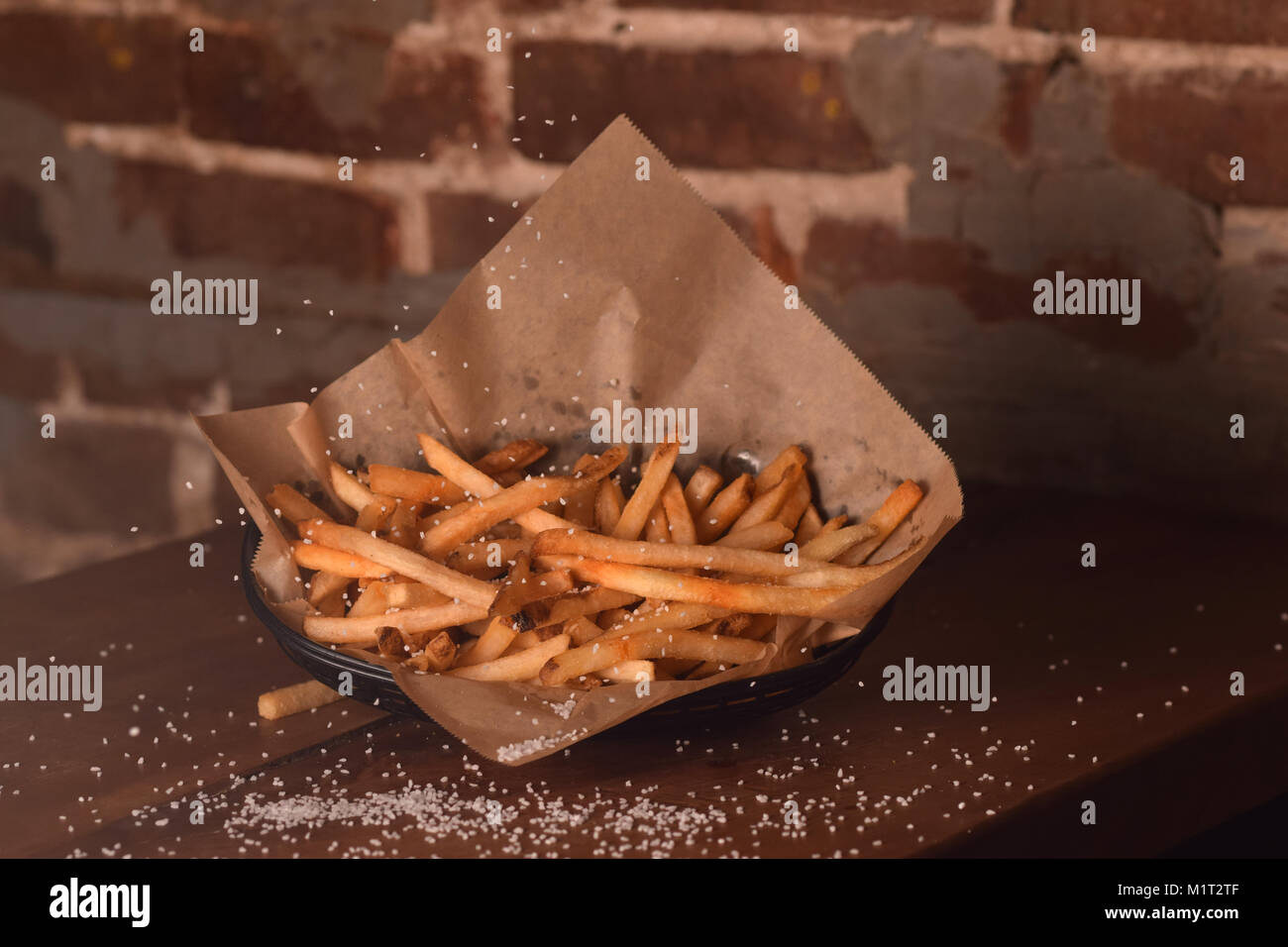 Golden brown French frieds with sprinkled salt on a wooden table against a brick wall. Stock Photo