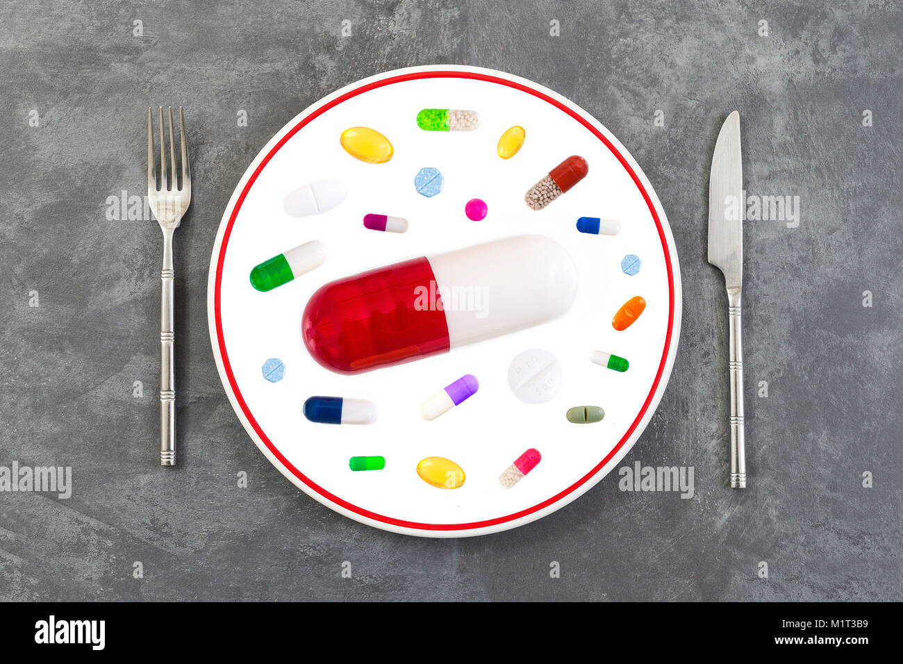 large sized red pill on a plate with silverware, in the middle of colorful medicine on grey slate background Stock Photo