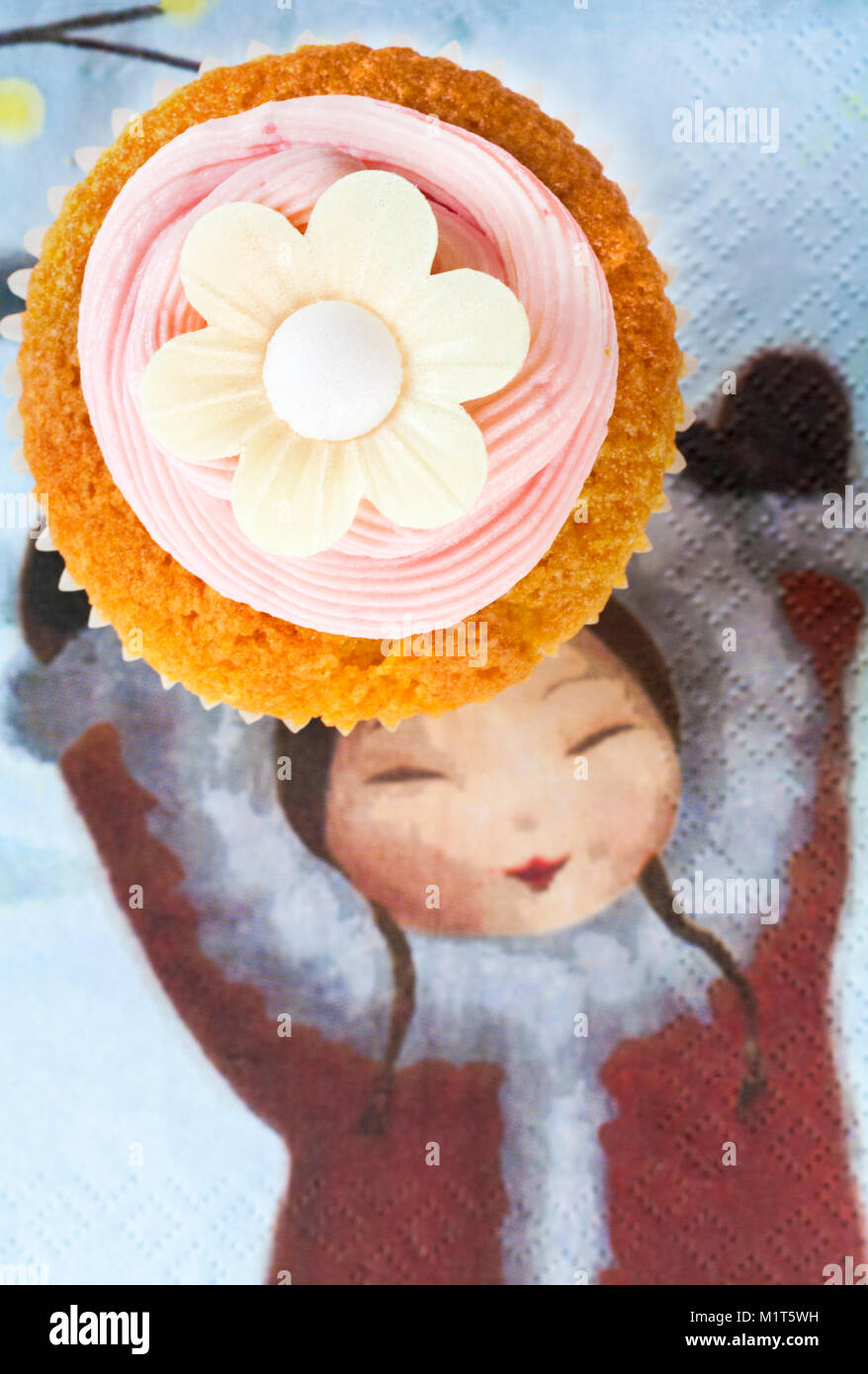 home made cupcake cake with strawberry pink frosting icing and rice paper edible daisy flower on top on serviette with girl holding hands up Stock Photo